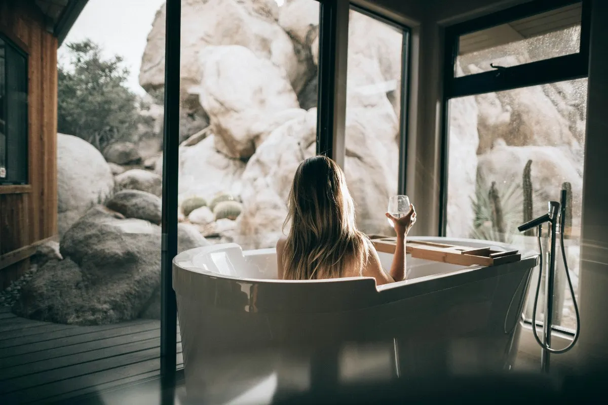 Cô gái tóc vàng cầm ly nước tắm bồn bên cửa sổ kính nhìn ra vách đá