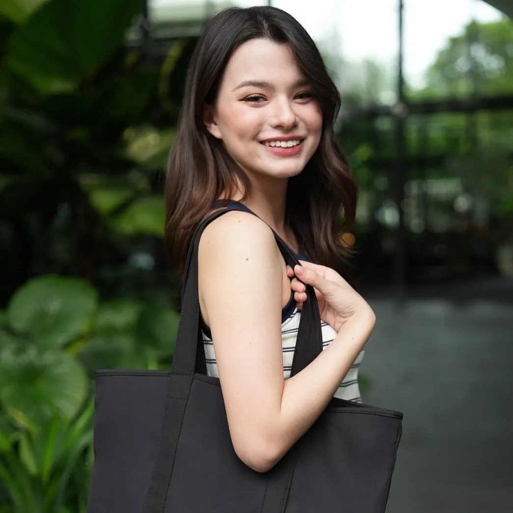 Cô gái trẻ rạng rỡ với nụ cười tươi, trang điểm nhẹ nhàng tông cam đào, kết hợp tóc uốn mềm và túi vải đen đơn giản, phù hợp với phong cách tự nhiên và năng động.