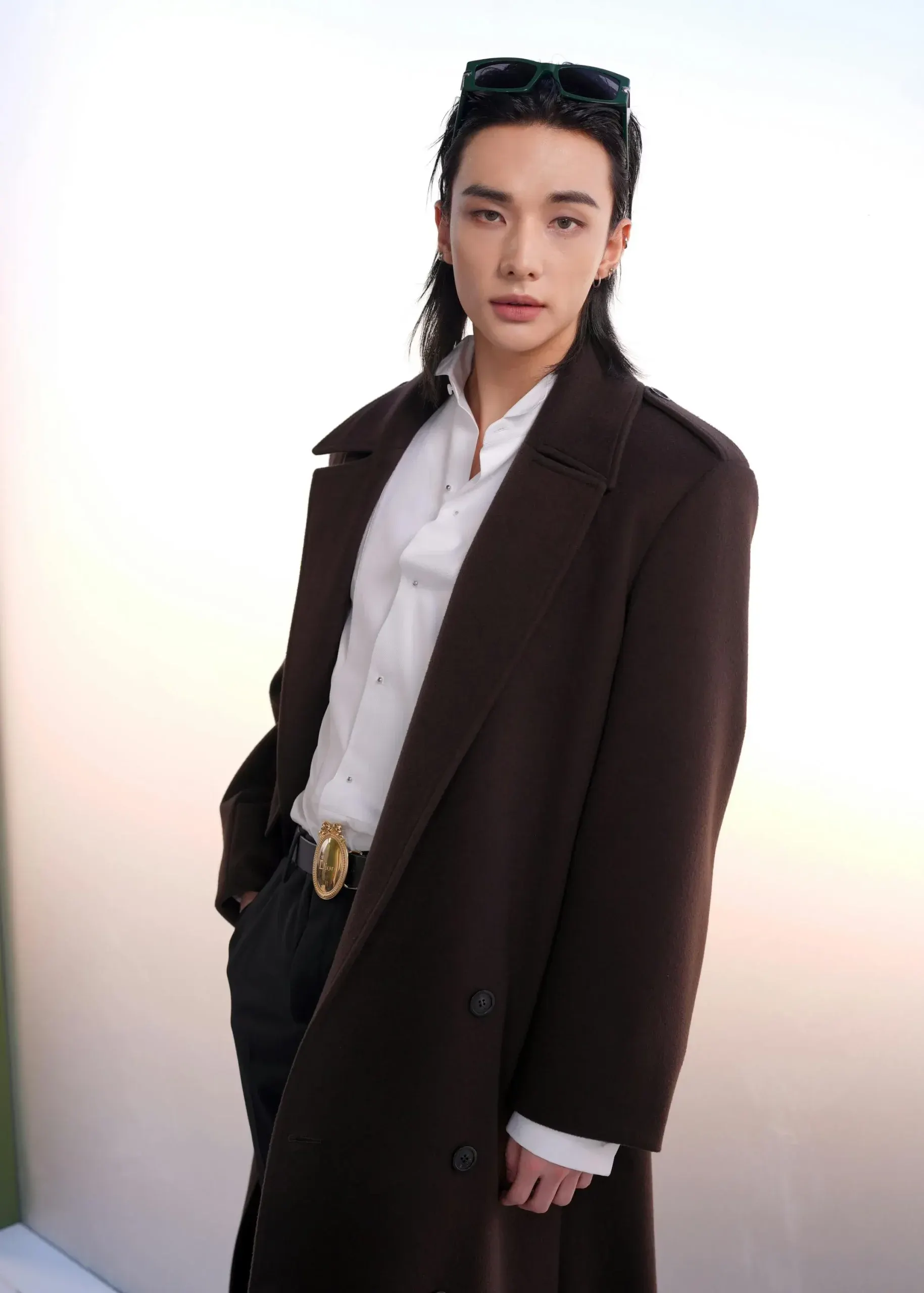 Hyunjin (Stray Kids) chọn áo khoác dáng dài màu nâu trầm mặc ngoài sơ mi trắng và quần tây đen, điểm nhấn nằm ở chiếc thắt lưng mặt vàng bản lớn.