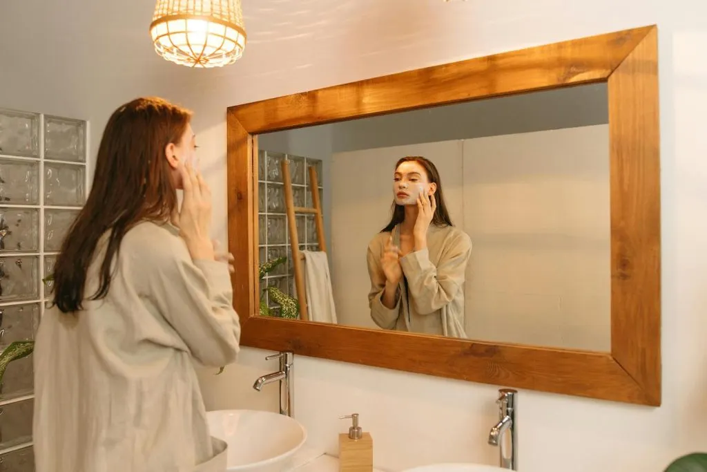 Cô gái trẻ đang đắp mặt nạ nghệ tự nhiên trong phòng tắm với khung gương gỗ, tạo không gian spa tại nhà ấm cúng