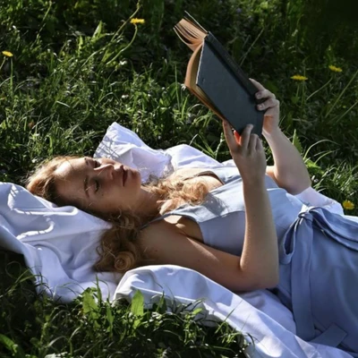 cô gái mặc đầm xanh tay cầm quyển sách nằm trên chiếc khăn trải trên bãi cỏ