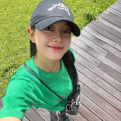 Cô gái đội mũ lưỡi trai đen, mặc áo thun xanh lá và đeo túi chéo, selfie ngoài trời với nụ cười rạng rỡ dưới ánh nắng, ngăn ngừa lão hóa