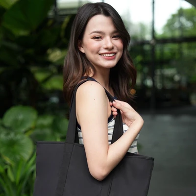 Cô gái trẻ rạng rỡ với nụ cười tươi, trang điểm nhẹ nhàng tông cam đào, kết hợp tóc uốn mềm và túi vải đen đơn giản, phù hợp với phong cách tự nhiên và năng động.