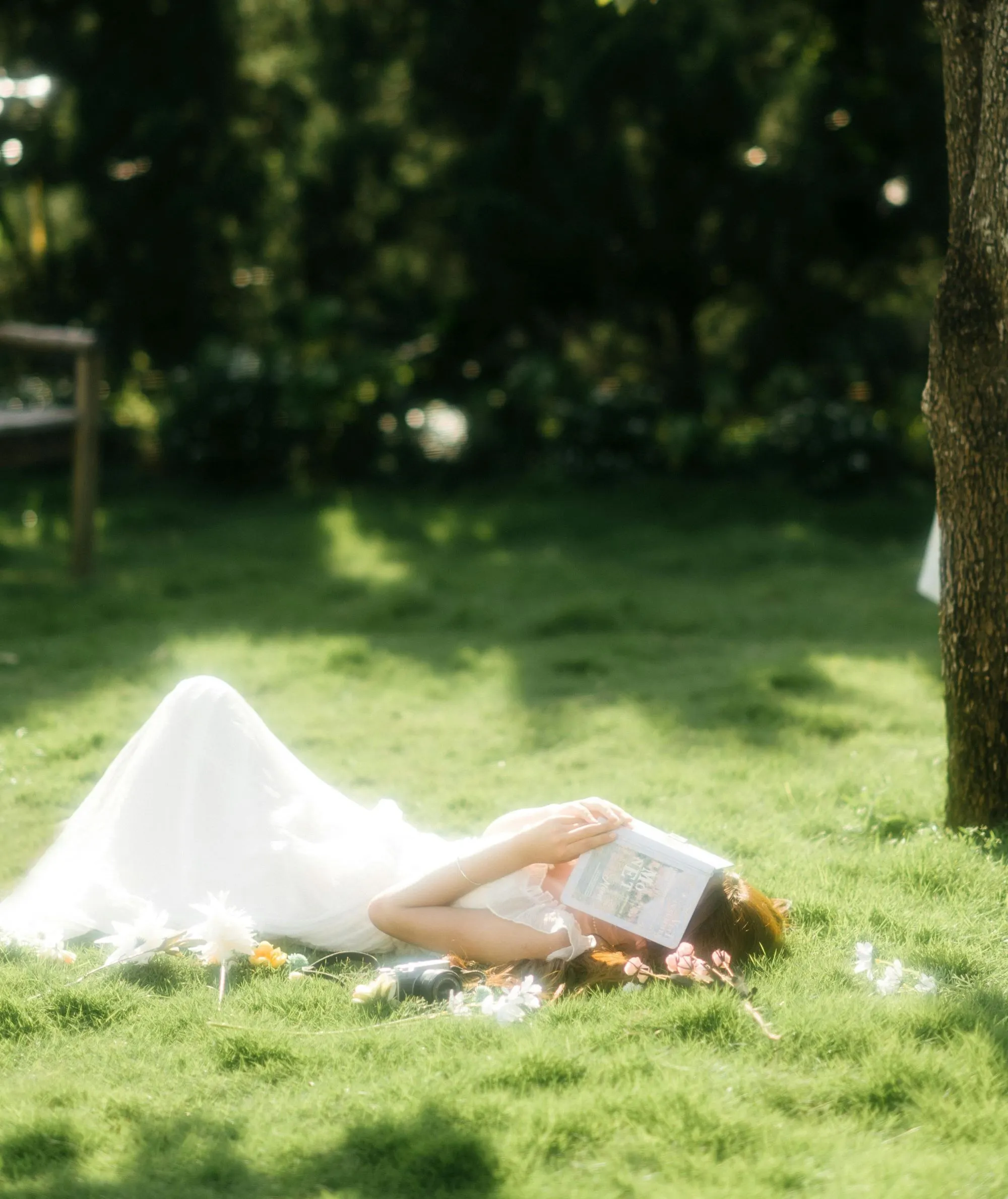 cô gái mặc đầm trắng nằm trên bãi cỏ xanh dưới ánh nắng mặt trời tay cầm quyển sách che mặt